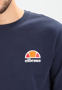 Sweat-shirt bleu marine en tissu doux, avec un logo elesse arrondi comportant des accents orange, rouge et blanc sur la poitrine. Col côtelé.