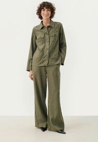 Completo due pezzi verde oliva composto da una camicia con bottoni e tasche sul petto, abbinata a pantaloni a gamba larga realizzati in tessuto testurizzato. Inclusi scarpe nere a punta.