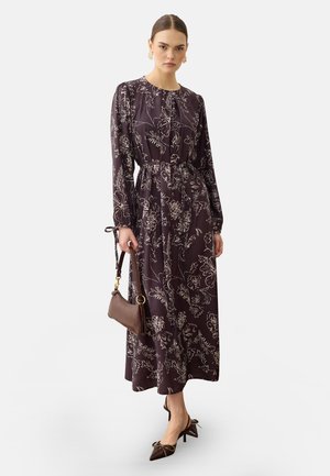 Femme portant une robe à manches longues à motif floral foncé, tenant un sac à main marron, assortie à des talons pointus marron et des boucles d'oreilles dorées, debout sur fond blanc.