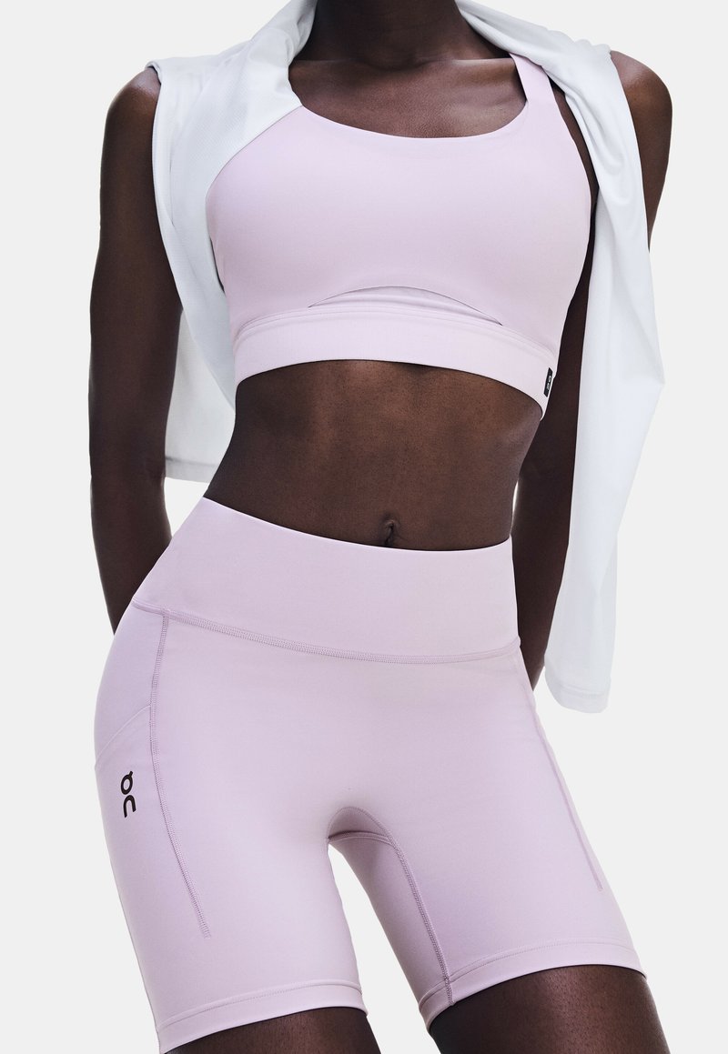 Person wearing light pink sports bra and matching high-waisted shorts with a white jacket draped over shoulders, standing against a plain background.