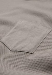 Camiseta de algodón gris con una superficie texturizada, que presenta un pequeño bolsillo de parche en el lado izquierdo del pecho. Cuello redondo simple y mangas cortas.