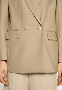 Blazer beige de doble botonadura con botones dorados, que presenta dos bolsillos frontales y una textura suave. Combinado con pantalones beige claros.