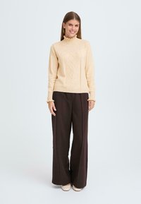 Un pull en tricot beige clair avec un col haut à volants, présentant un motif en losanges texturé, associé à un pantalon large marron foncé.