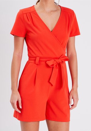 Kvinde iført kortærmet rød playsuit med V-hals, læg ved taljen og et stofbælte bundet i en sløjfe foran.
