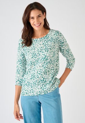 Frau lächelt, trägt ein langärmliges Hemd mit weiß-grünem Muster und hellblaue Jeans, steht mit einer Hand in der Tasche vor einem weißen Hintergrund.