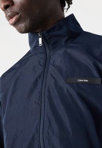 Veste légère bleu marine avec un col montant et une fermeture éclair. Présente un patch logo de marque noir sur la poitrine. Texture lisse.