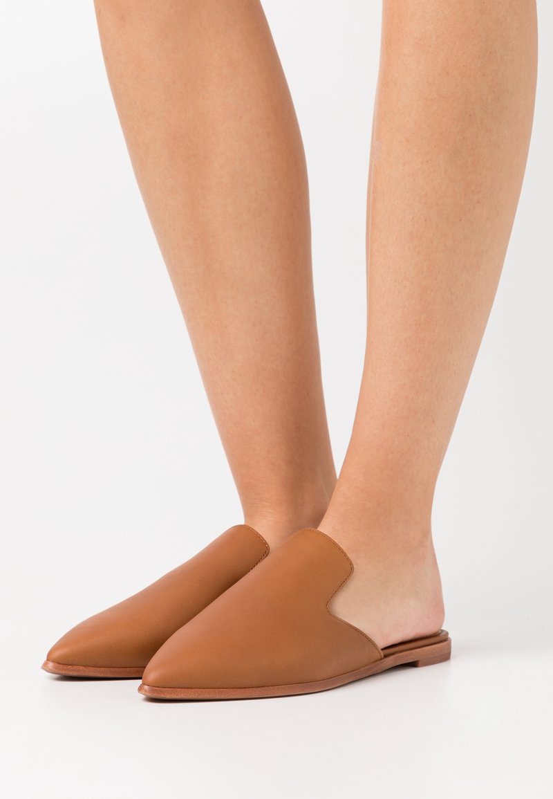 Pieds nus portant des mules en cuir marron clair à enfiler, avec des bouts pointus, présentés sur un fond blanc uni.