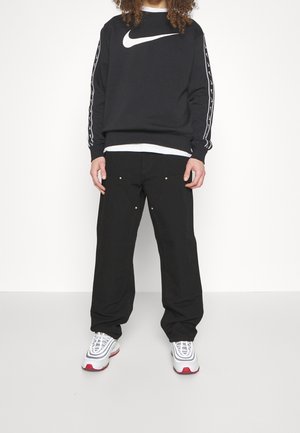 Person trägt schwarzen Nike-Sweatshirt mit weißem Swoosh, schwarze Hose und weiße Sneakers mit roten Sohlen, steht vor weißem Hintergrund.