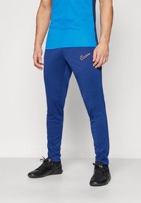 Blaue Sporthose aus Stretchmaterial, mit schlanker Passform, orangefarbenem Nike-Logo und elastischen Bündchen. Getragen mit schwarzen Turnschuhen.