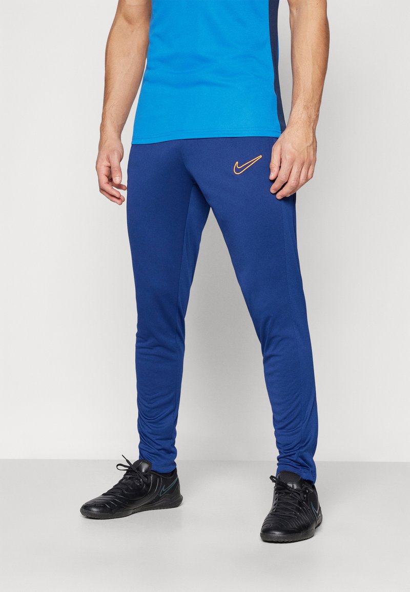 Blaue Sporthose aus Stretchmaterial, mit schlanker Passform, orangefarbenem Nike-Logo und elastischen Bündchen. Getragen mit schwarzen Turnschuhen.
