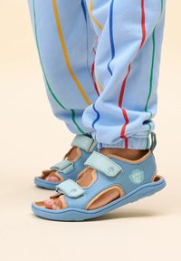 Sandalias de color azul claro con dos correas ajustables, de cuero suave y suelas de goma; combinadas con pantalones azules a juego que presentan rayas coloridas.