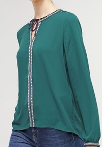 Kvinna i en turkos blus med lång ärm, mönstrad kant och knytning vid halsen, tillsammans med blå denimjeans.