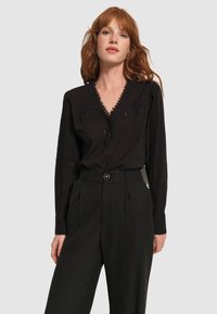 Blusa nera a maniche lunghe con scollatura profonda, bordi in pizzo e ricami, abbinata a pantaloni neri a vita alta.