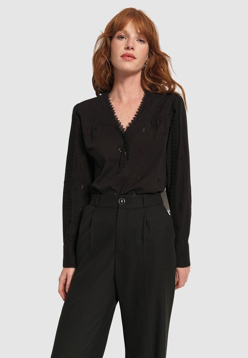 Blusa nera a maniche lunghe con scollatura profonda, bordi in pizzo e ricami, abbinata a pantaloni neri a vita alta.