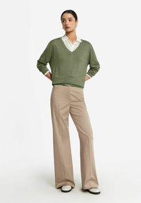 Zielony sweter w serek z miękkiej dzianiny, noszony na białej koszuli z kołnierzykiem. Jasnobeżowe spodnie o szerokich nogawkach i białe buty z czarną podeszwą.