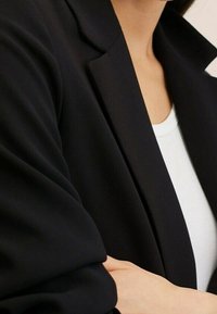 Blazer noir avec revers structurés, texture de tissu douce, manches ajustées, porté par-dessus un haut blanc, montrant un gros plan du décolleté.