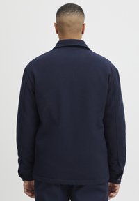 Jachetă dintr-un amestec de lână de culoare bleumarin, cu guler tăiat și manșete cu buton, având o textură netedă și linii curate într-un design relaxat.