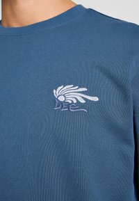 Camisa de algodón azul de manga larga con un acabado texturizado y un logo bordado en blanco en el lado izquierdo del pecho. Escote redondo.