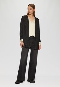 Cardigan noir ample avec des paillettes discrètes sur un chemisier crème. Des jeans noirs taille haute à jambes larges et des bottes noires imposantes complètent le look.