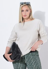 Blonde Frau, die ein weißes Langarmshirt und gemusterte Hosen trägt, hält eine große schwarze Leder-Clutch in der Hand und hat schwarze Sonnenbrillen auf dem Kopf.