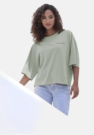 Locker sitzendes, kurzärmliges T-Shirt aus hellgrüner Baumwolle mit rundem Halsausschnitt und dezentem Branding. Kombiniert mit hellblauen Jeans.