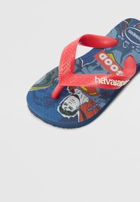 Rubber flip-flop with red straps and a blue sole featuring comic-style graphics of a hero, with textured footbed for grip.
