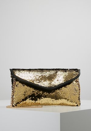 Pochette enveloppe dorée à sequins avec bordure noire et bandoulière chaîne dorée, posée sur une surface d'exposition blanche sur fond gris.