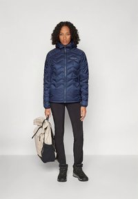 Tumesinine tikitud puffer-jakk lukuga ees, chevron-mustriga, kantud mustade pükste ja mustade matkasaabastega, hoides käes beeži ja musta seljakotti.