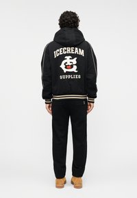 Μαύρο φούτερ με κουκούλα με το κείμενο "ICECREAM SUPPLIES" και γραφικό στην πλάτη, συνδυασμένο με μαύρο παντελόνι και ταμπά μποτάκια. Ριμπ μανσέτες και τελείωμα.