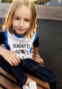 T-Shirt bianco con accenti blu e maniche nere raffigurante il testo "BUGATTI". Jeansu scuri con cuciture visibili e sneaker bianche.