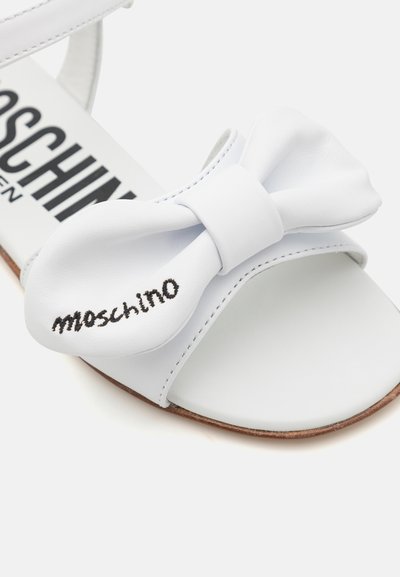 Bílé kožené sandály s velkou mašlí a vyraženým logem "Moschino". Hladký povrch, otevřená špička a hnědá podrážka.