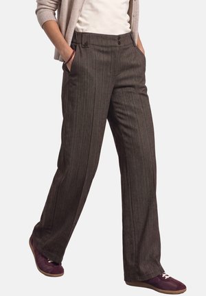 Personne portant un pantalon large marron à fines rayures, un haut blanc, un cardigan beige et des baskets violettes, les mains dans les poches, debout devant un fond blanc.