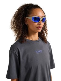 Vrouw met krullend haar met een beige montuur en blauwe gespiegelde zonnebril en een donkergrijs T-shirt met "HALO" op de borst gedrukt.