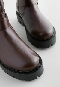 Botas de cuero marrón hasta el tobillo con un acabado texturizado, suelas de goma negra gruesa con surcos profundos y costuras reforzadas a lo largo de los bordes.