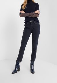 Jeans negros de talle alto con corte recto y dobladillo sin terminar, combinados con un suéter de cuello alto azul marino con mangas de patrón y botas de tobillo azul marino brillantes.