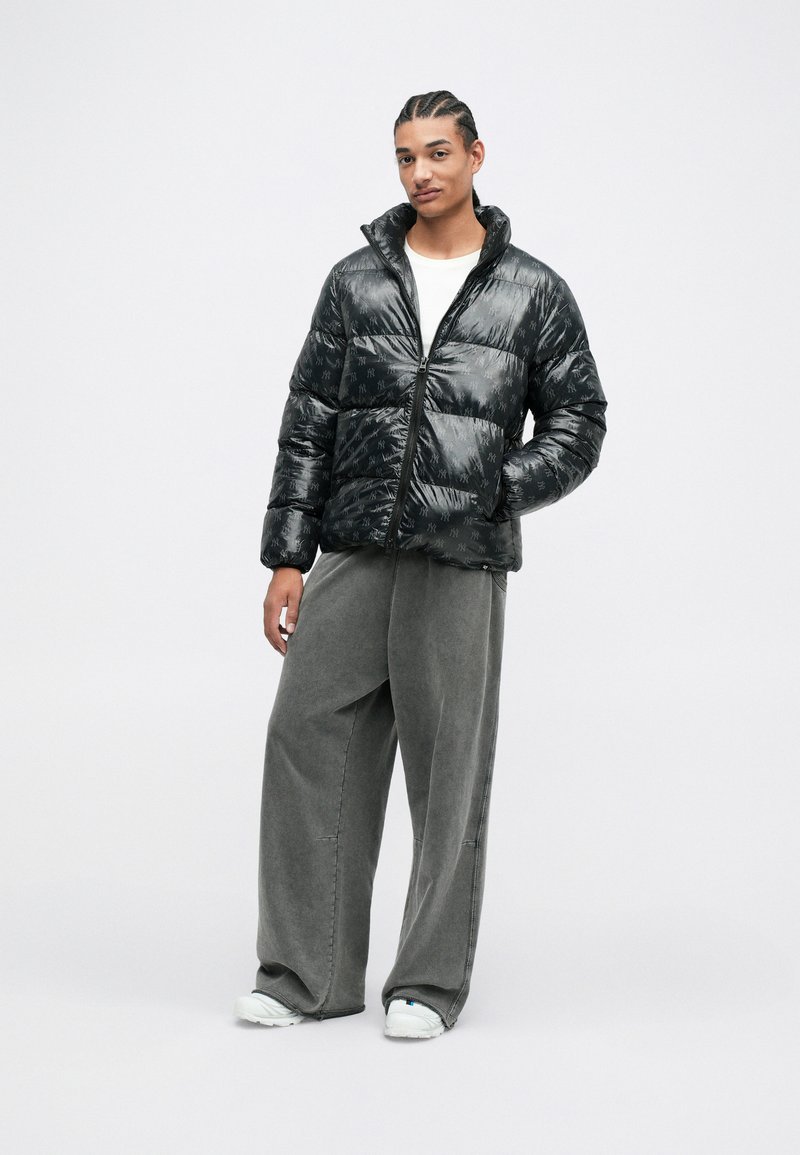 Schwarze Pufferjacke mit glänzender Oberfläche, Reißverschluss und Stehkragen. Über grauen weit geschnittenen Hosen und weißen Schuhen getragen.