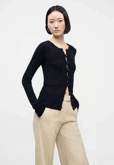 Kvinne med kort svart hår iført en svart knappet cardigan og beige bukser, står med hendene i lommene mot en ensfarget bakgrunn.