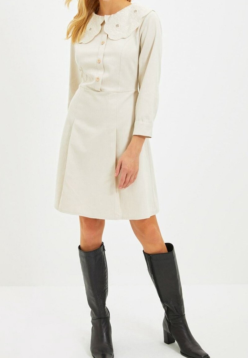 Vestido de color crema con un gran collar bordado y botonadura frontal, con mangas largas y cintura ajustada. Combinado con botas negras hasta la rodilla.