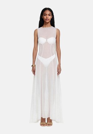 Donna con capelli lunghi che indossa un abito lungo bianco trasparente senza maniche sopra reggiseno e slip bianchi, braccialetto d’oro e tacchi aperti dorati.