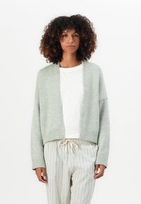 Vrouw met krullend haar, gekleed in een lichtgrijze open cardigan, witte top en beige gestreepte broek met trekkoord, staande voor een witte achtergrond.