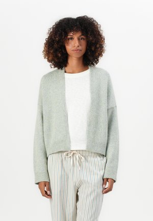 Kvinne med krøllete hår iført en lys grå åpen cardigan, hvit topp og beige stripete bukser med snøring, stående mot en hvit bakgrunn.