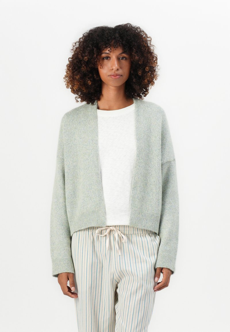 Vrouw met krullend haar, gekleed in een lichtgrijze open cardigan, witte top en beige gestreepte broek met trekkoord, staande voor een witte achtergrond.