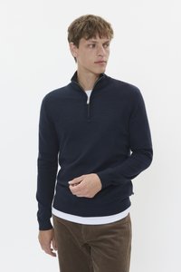 Navy blue zip-neck sweater in a soft, textured knit. Fitted design with ribbed cuffs, worn over a white shirt, paired with corduroy trousers.