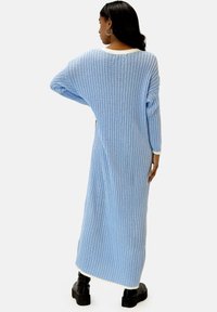 Blauwe ribgebreide jurk met lange mouwen en een brede halslijn, voorzien van een witte bies bij de kraag en zoom. Draagt zwarte schoenen.