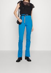 Haut en dentelle noire avec des manches à volants, associé à un pantalon ajusté bleu avec une ceinture. Le mannequin porte des bottes à talons noires et tient un petit sac.