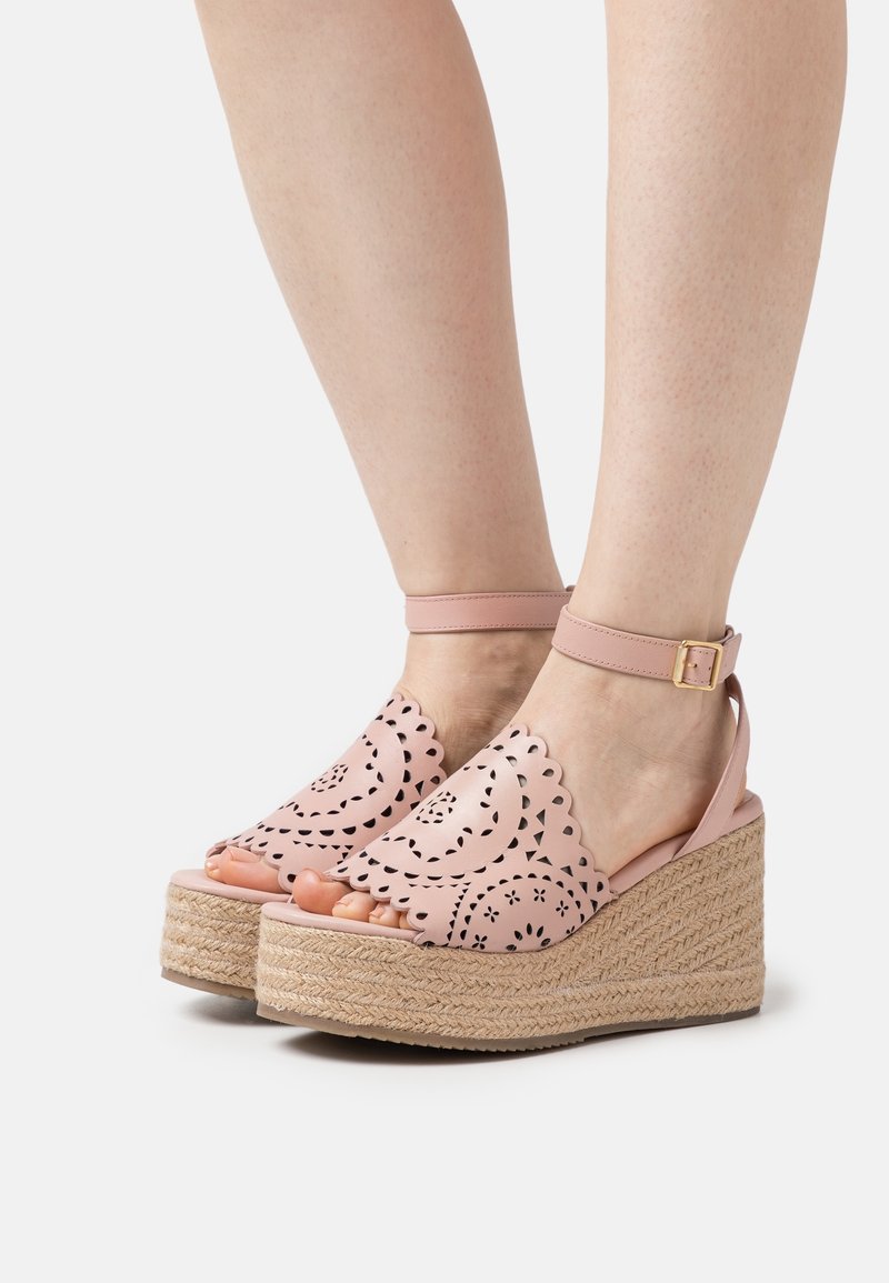 Jambes portant des sandales compensées à bride à la cheville de couleur rose clair, ornées de motifs décoratifs festonnés et découpés au laser, avec des semelles plateformes en corde tressée.