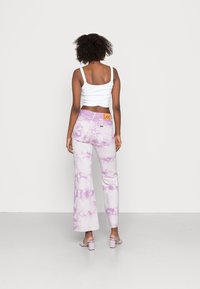 Jeans flare em tie-dye roxo com uma textura suave, apresentando uma cintura com marca e bolsos traseiros, combinados com sandálias de bloco lavanda.
