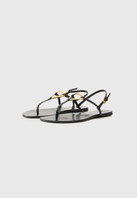 Sandali neri piatti con sottili cinturini incrociati. Dettagli in metallo dorato che accentuano la parte anteriore. Design della suola liscio e minimalista.