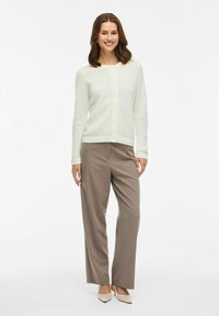Cárdigan de punto en un suave color crema con un patrón texturizado, cierre de botones y puños acanalados, combinado con pantalones marrón claro y tacones beige.