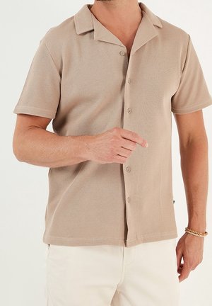 Korte mouwen, knoopshirt in lichtbeige, met een geribbelde textuur en een klassieke kraag. De stof is zacht en lichtgewicht.